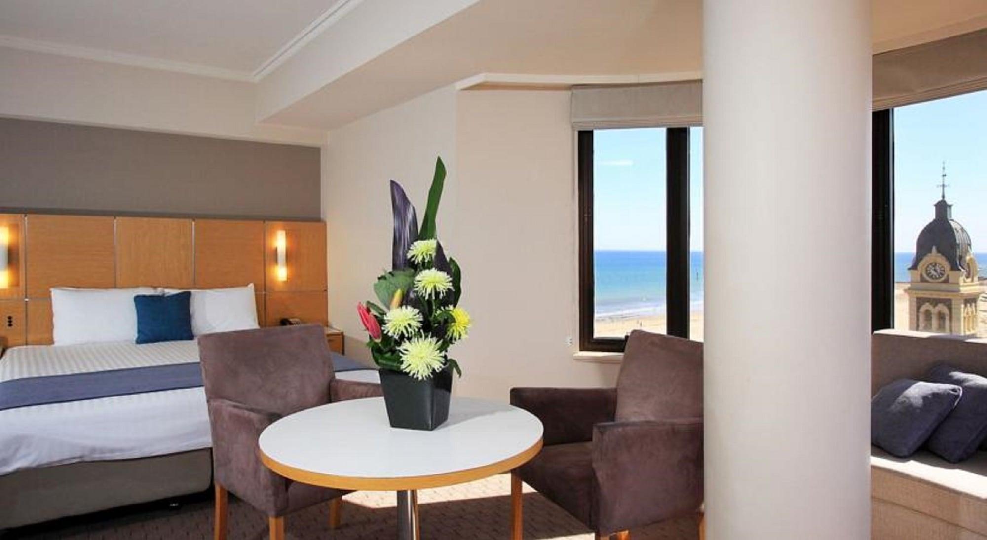 Hotel Stamford Grand Adelaide Glenelg Exterior foto The photo shows a cozy hotel room with a comfortable seating arrangement. There are two brown chairs positioned around a round table, which features a decorative flower arrangement in a black vase. The background includes a window that offers a view 