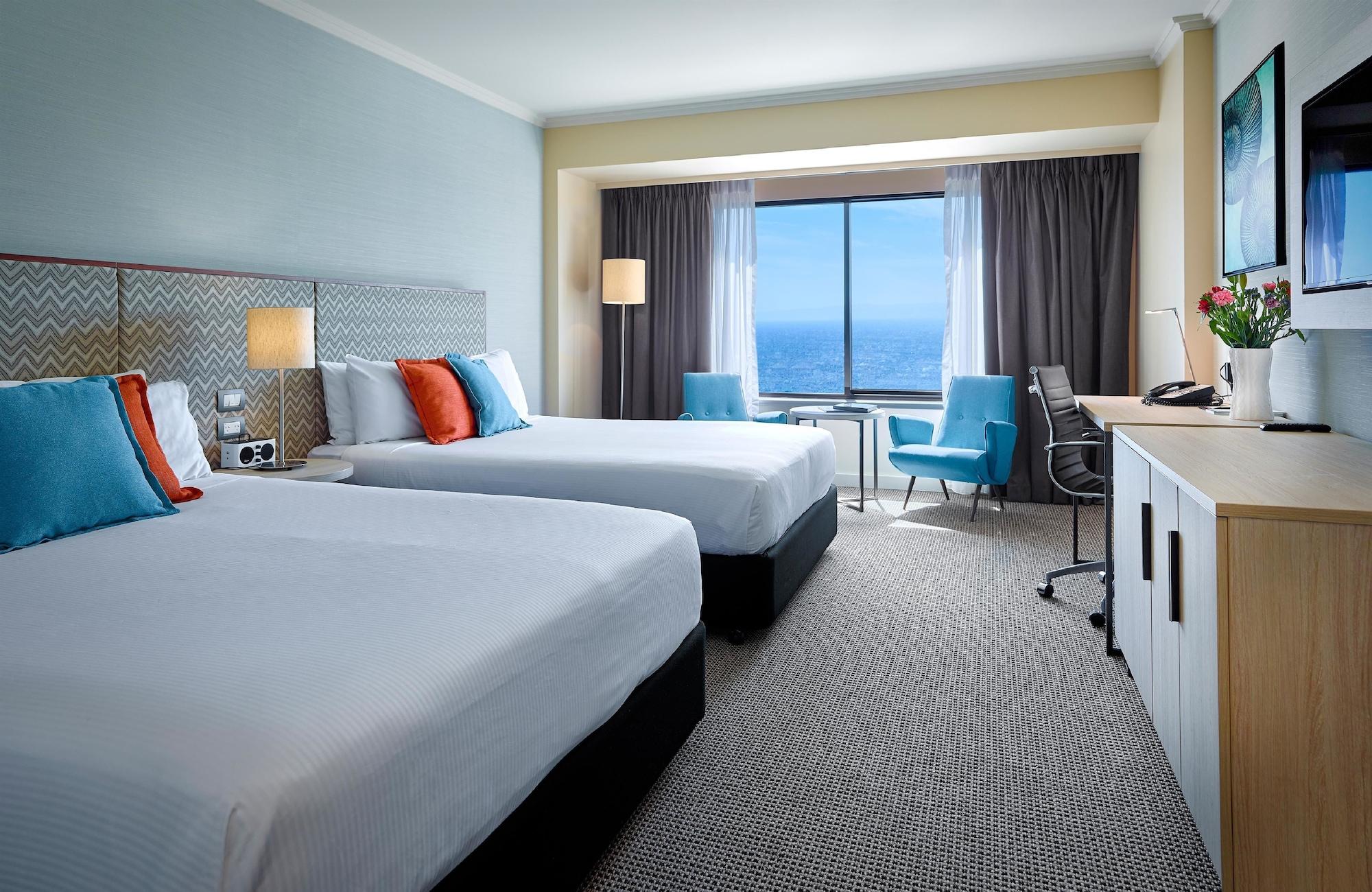 Hotel Stamford Grand Adelaide Glenelg Exterior foto The photo shows a well-decorated hotel room featuring two queen-sized beds with white linens and colorful accent pillows. There is a large window with curtains that offers a view of the ocean. A small seating area with a blue chair and a matching des