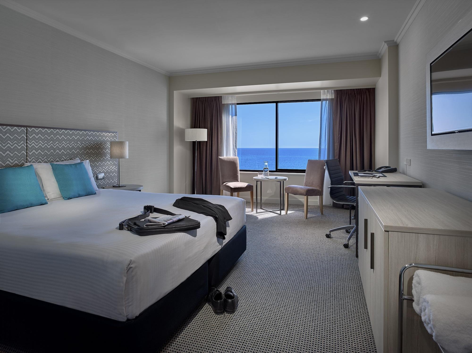 Hotel Stamford Grand Adelaide Glenelg Exterior foto The photo shows a modern hotel room featuring a queen-sized bed with white linens and two aqua blue decorative pillows. There are bedside lamps and a large window with a view of the ocean. The room also has a small seating area with two chairs and a 