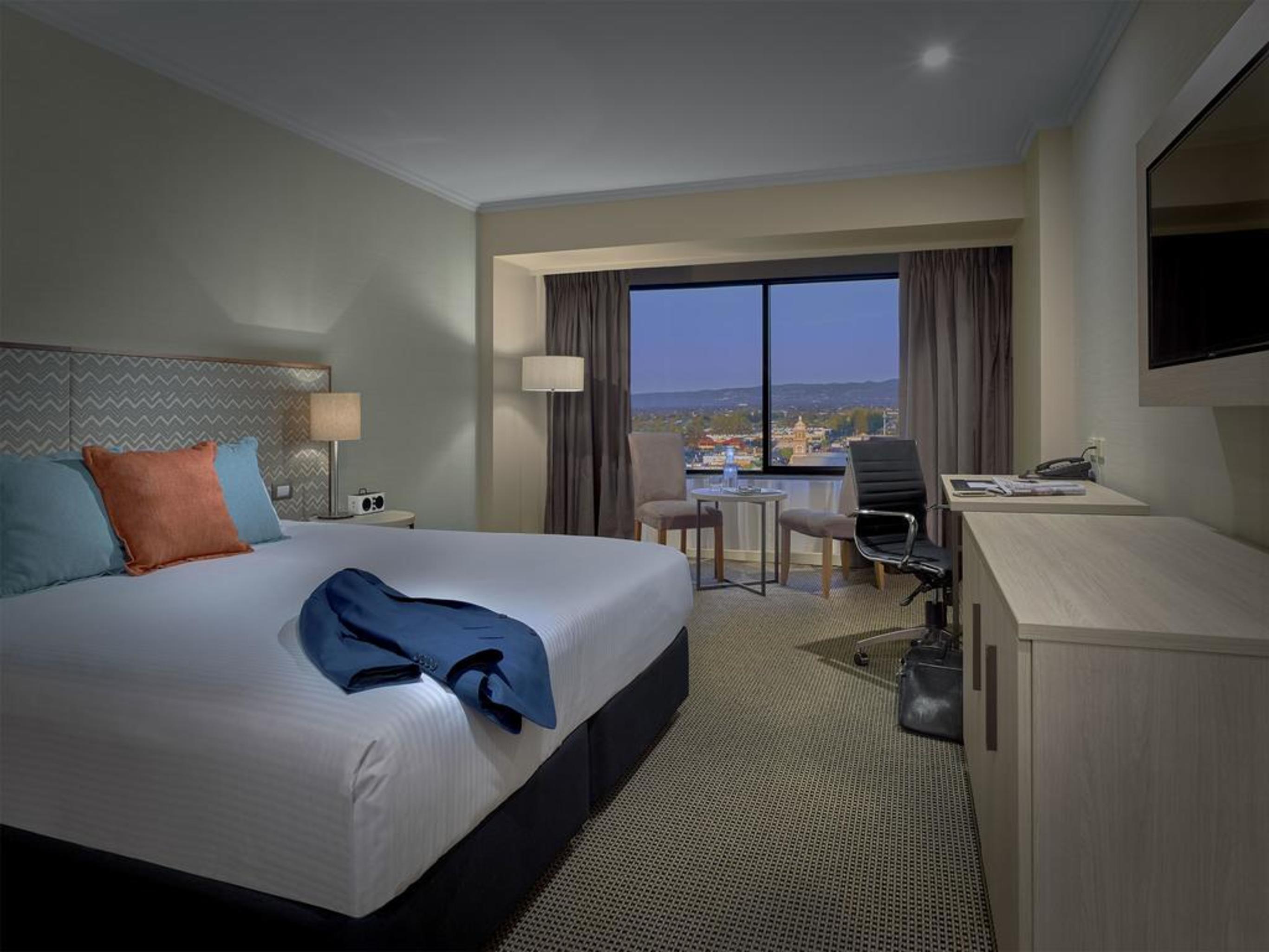 Hotel Stamford Grand Adelaide Glenelg Exterior foto The photo shows a modern hotel room. It features a large double bed with white bedding and decorative pillows in orange and blue. There's a nightstand with lamps on either side of the bed. A large window overlooks a view outside, and there's a desk a