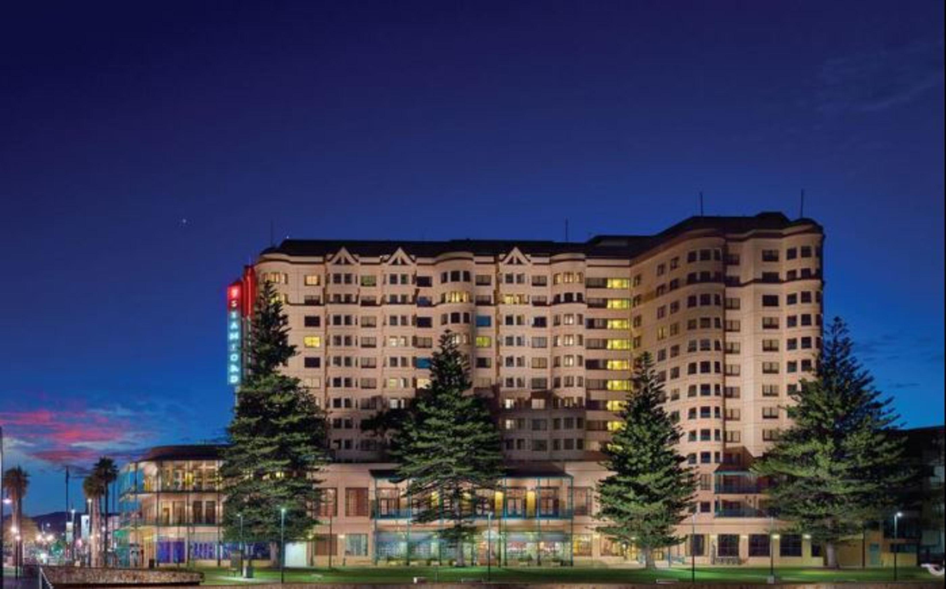 Hotel Stamford Grand Adelaide Glenelg Exterior foto The photo displays a multi-story hotel building at dusk, illuminated by various lights. The architecture features a mix of modern and traditional design elements, with numerous windows showcasing a warm glow. Surrounding the hotel are several tall tr