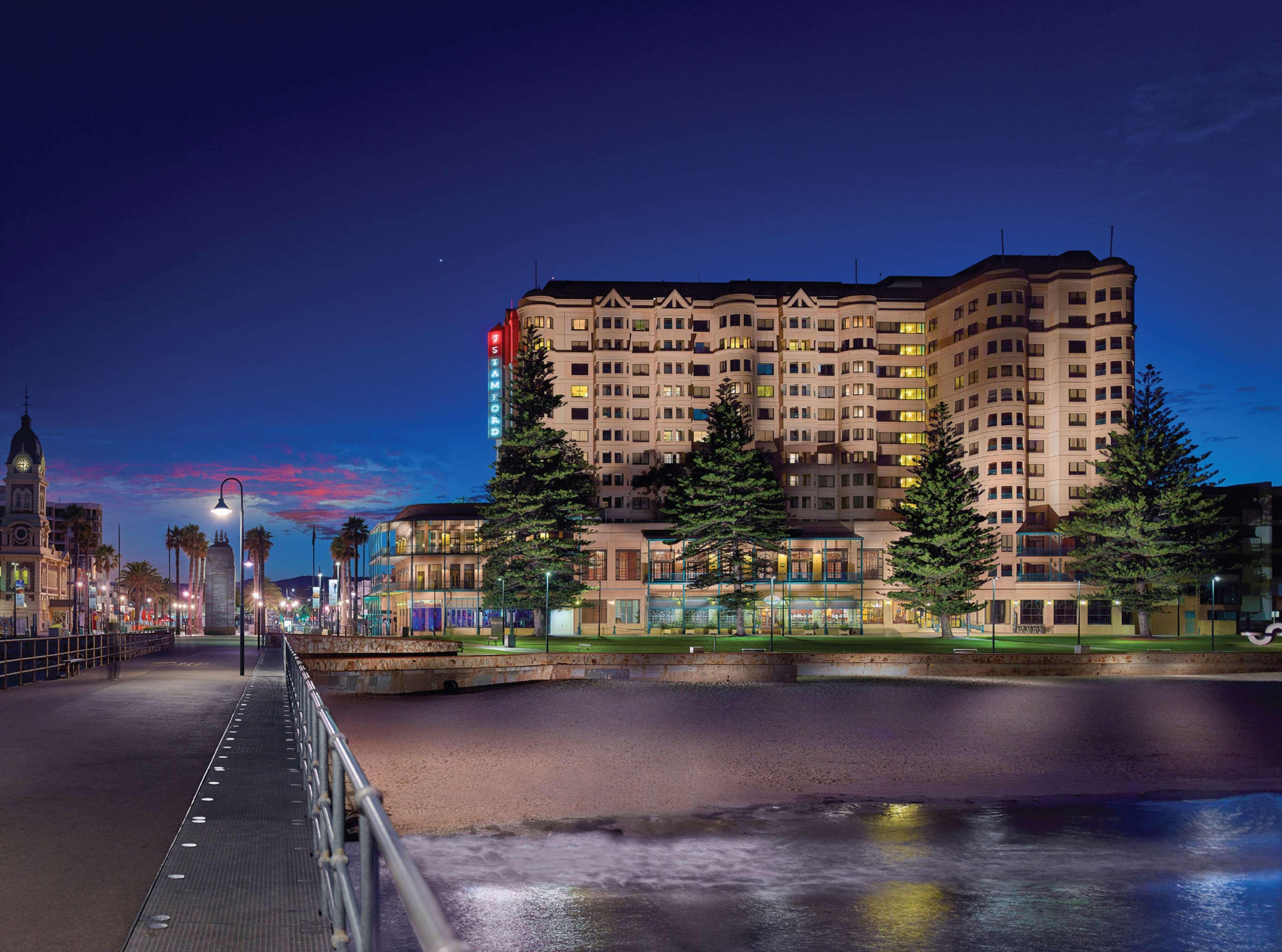 Hotel Stamford Grand Adelaide Glenelg Exterior foto The photo shows a waterfront scene at dusk, featuring a large building lit up against a darkening sky. The architecture appears modern and prominent, with several windows illuminated. In the foreground, there is a walkway or pier with fencing along t