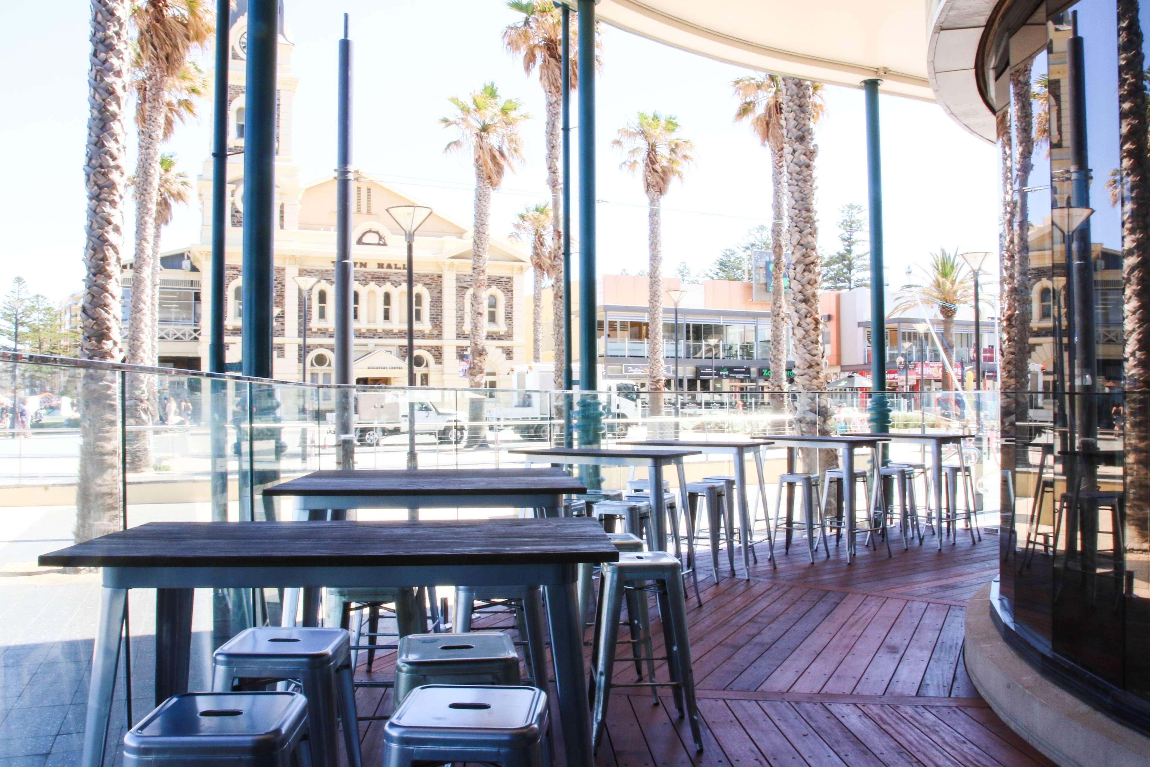 Hotel Stamford Grand Adelaide Glenelg Exterior foto The photo shows an outdoor seating area of a café or restaurant. It features metal tables and stools arranged on a wooden deck. The setting has large glass panels, providing an open and airy atmosphere. In the background, palm trees and a building wi