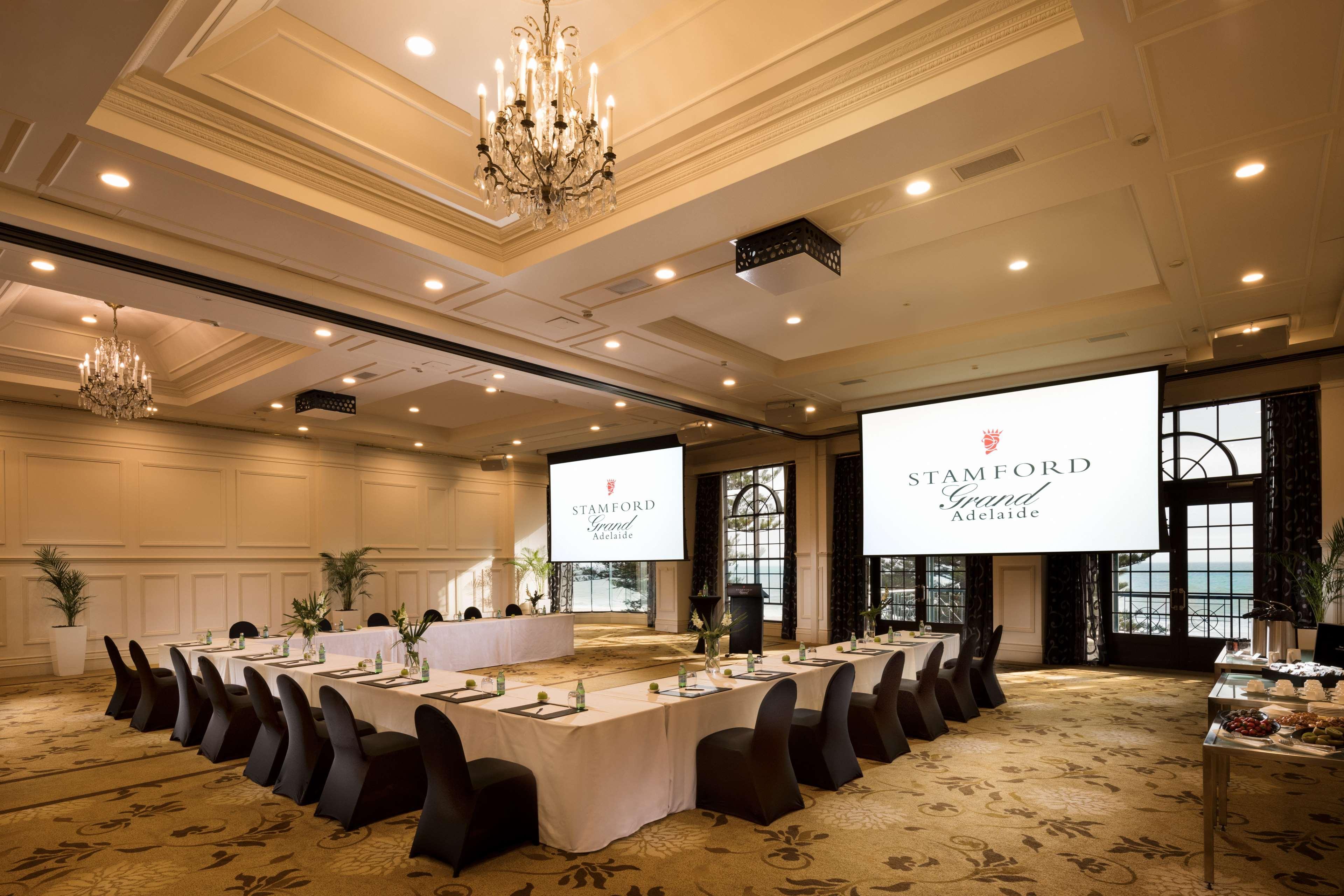 Hotel Stamford Grand Adelaide Glenelg Exterior foto The photo shows a spacious and elegantly designed conference room. The room features a long table set for a meeting, surrounded by stylish chairs. Two large screens are positioned at the front, displaying the logo "Stamford Grand Melbourne." The ceil