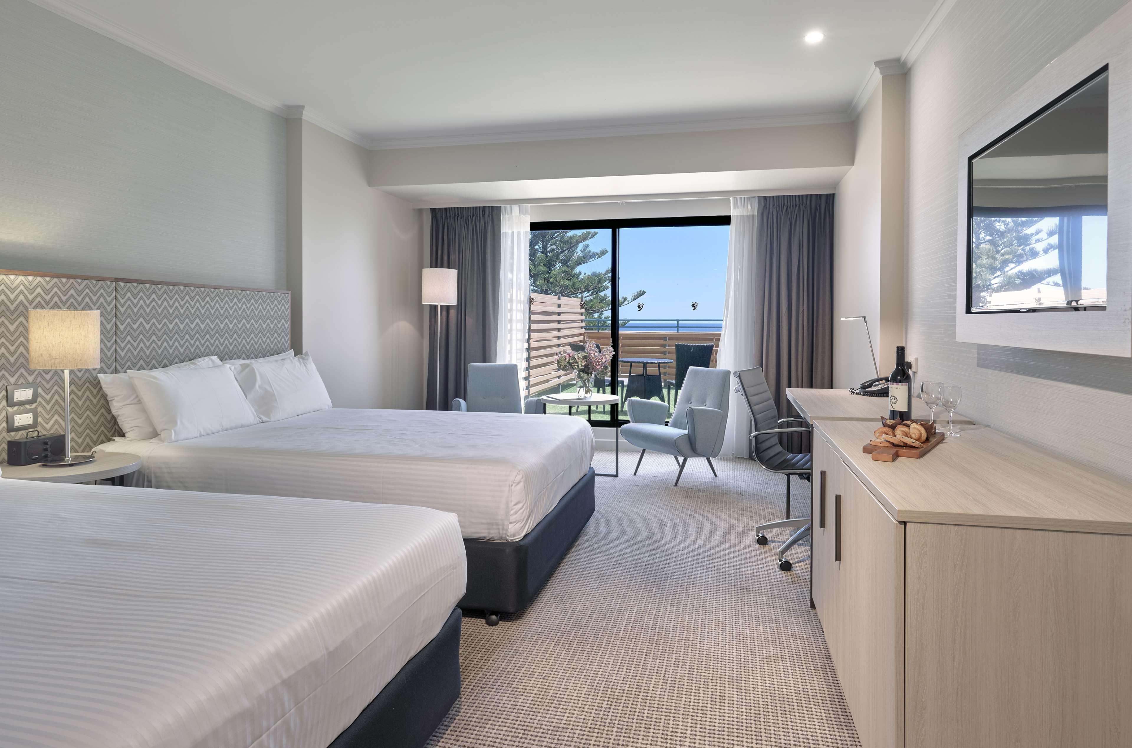 Hotel Stamford Grand Adelaide Glenelg Exterior foto The photo shows a modern hotel room featuring two beds with white linens. There's a large window that provides natural light and a view of a balcony area. The room includes two chairs and a small table, creating a cozy sitting area. A television is m