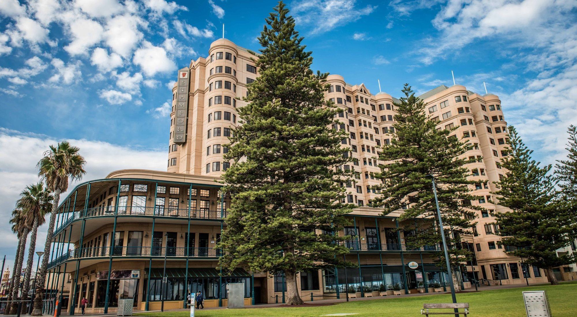 Hotel Stamford Grand Adelaide Glenelg Exterior foto The image shows a large building with a distinctive architectural style, characterized by a tan or beige color. The structure appears to have multiple floors and rounded edges. In front of the building, there are several tall green trees, which add t